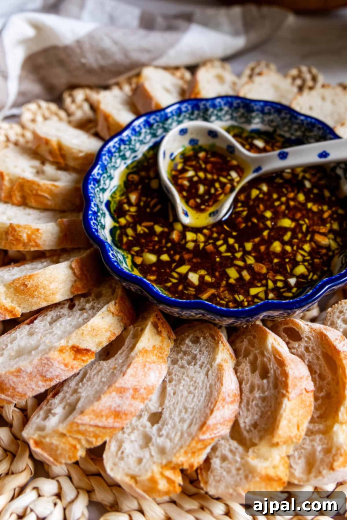 A bowl of oil and herb dip with a hint of balsamic vinaigrette sits with a spoon, surrounded by slices of crusty bread arranged in a circular pattern on a woven mat.