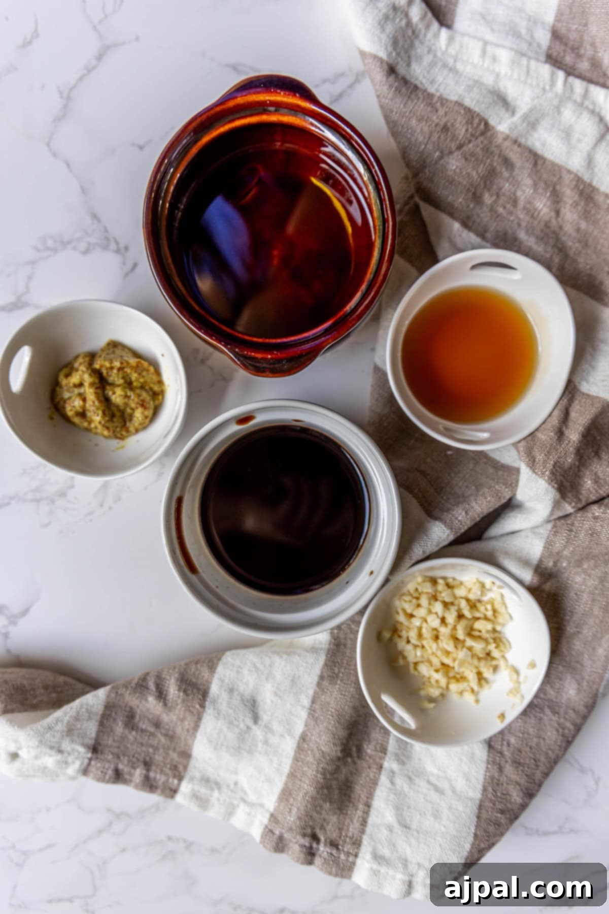 Five small bowls on a striped cloth contain ingredients for a vinaigrette: one with brown liquid, one with mustard, one with light brown liquid, one with chopped garlic, and one with dark balsamic.