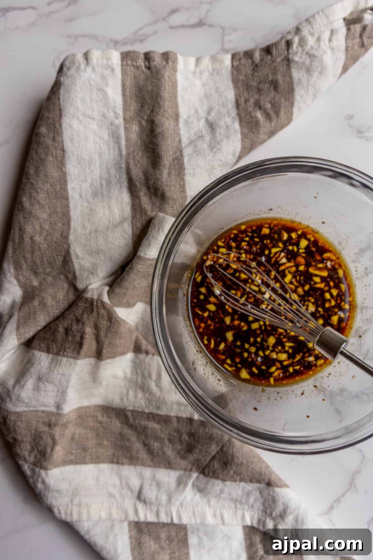 A glass bowl with a metal whisk mixing a dark balsamic vinaigrette with chopped ingredients, placed on a beige and white striped cloth on a marble surface.