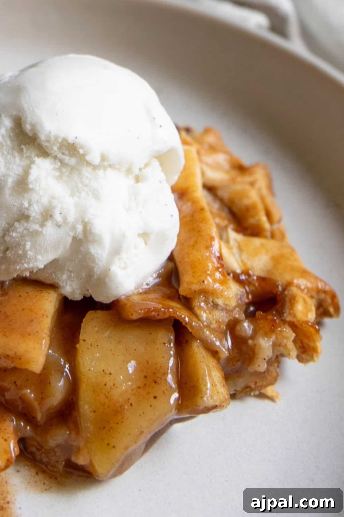 Heirloom Apple Pie 2 A slice of classic apple pie is topped with a scoop of vanilla ice cream, served on a white plate for the perfect comforting treat.