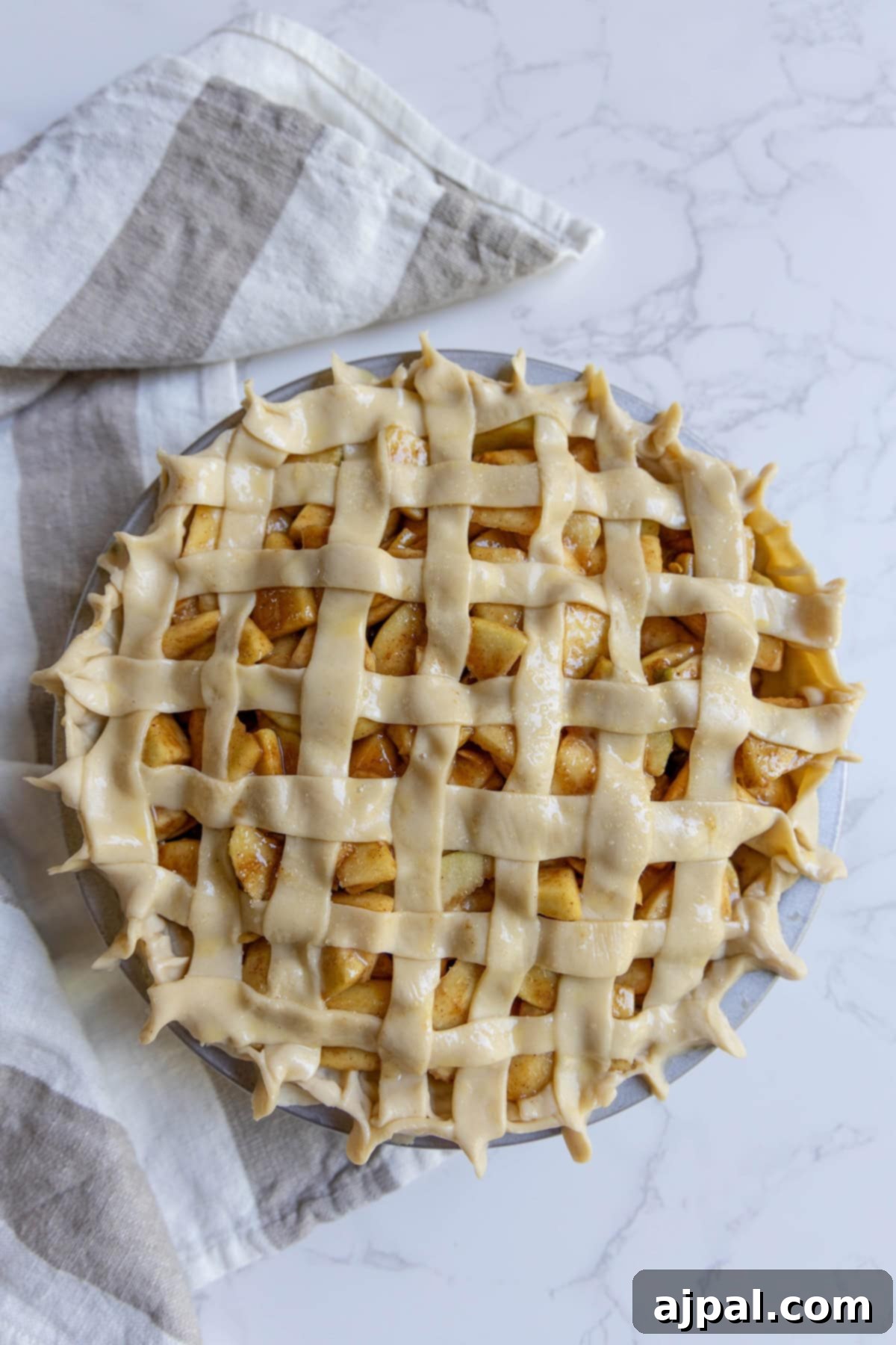 Heirloom Apple Pie 11 Unbaked apple pie with a lattice crust sits on a marble surface, next to a striped kitchen towel, ready to become a classic homemade apple pie.