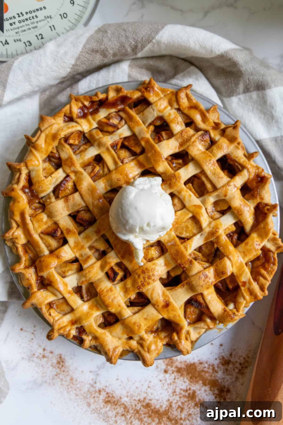 Heirloom Apple Pie 12 A classic baked apple pie with a lattice crust, topped with vanilla ice cream, rests on a striped kitchen towel, accompanied by a scale and a dusting of cinnamon.