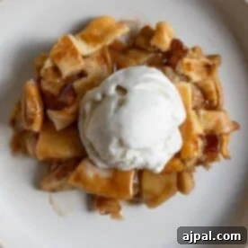 A slice of apple pie with pieces of crust is topped with a scoop of vanilla ice cream on a white plate.