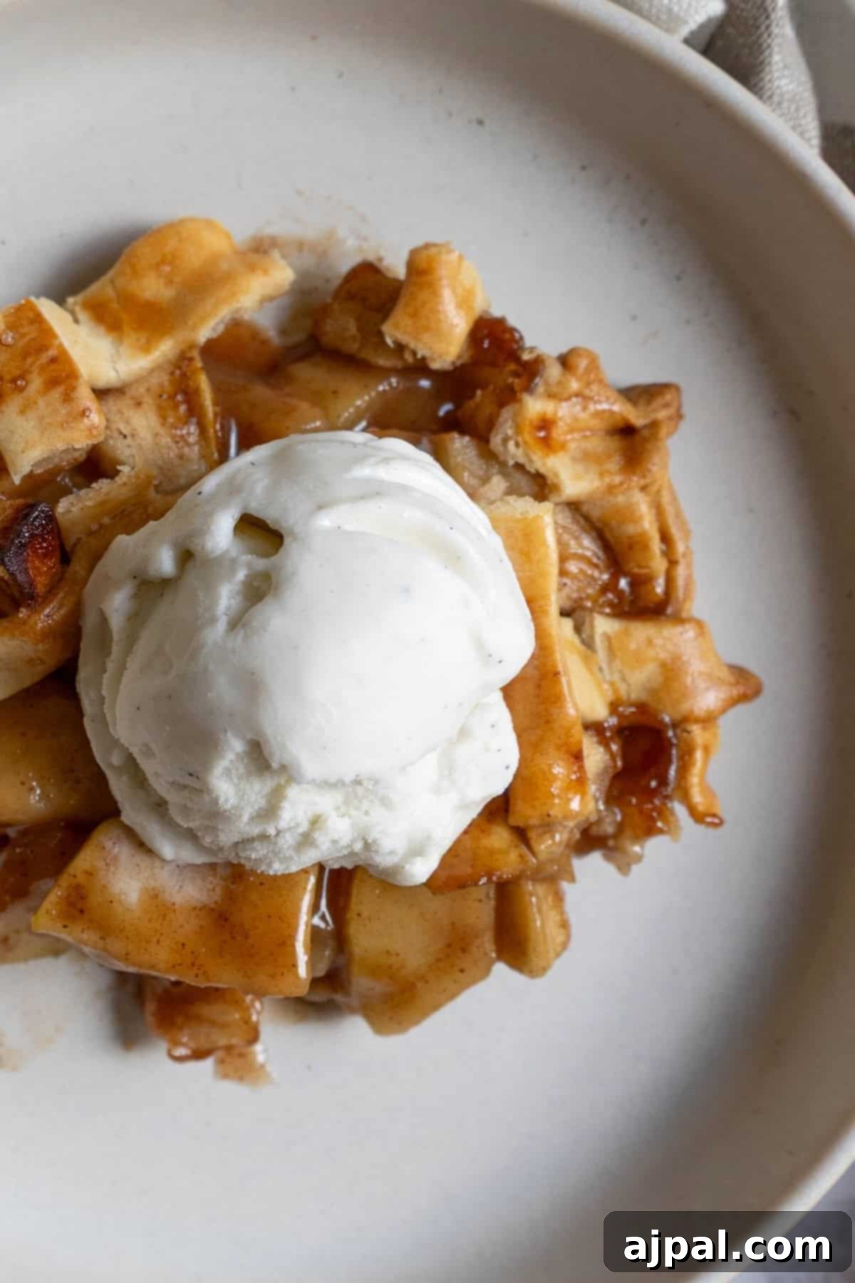 Heirloom Apple Pie 3 A slice of apple pie topped with a scoop of vanilla ice cream rests on a beige plate, creating the perfect classic dessert.