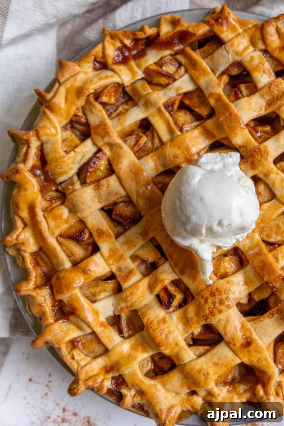 Heirloom Apple Pie 4 A classic apple pie with a golden lattice top and a scoop of vanilla ice cream, beautifully displayed on a light-colored surface.