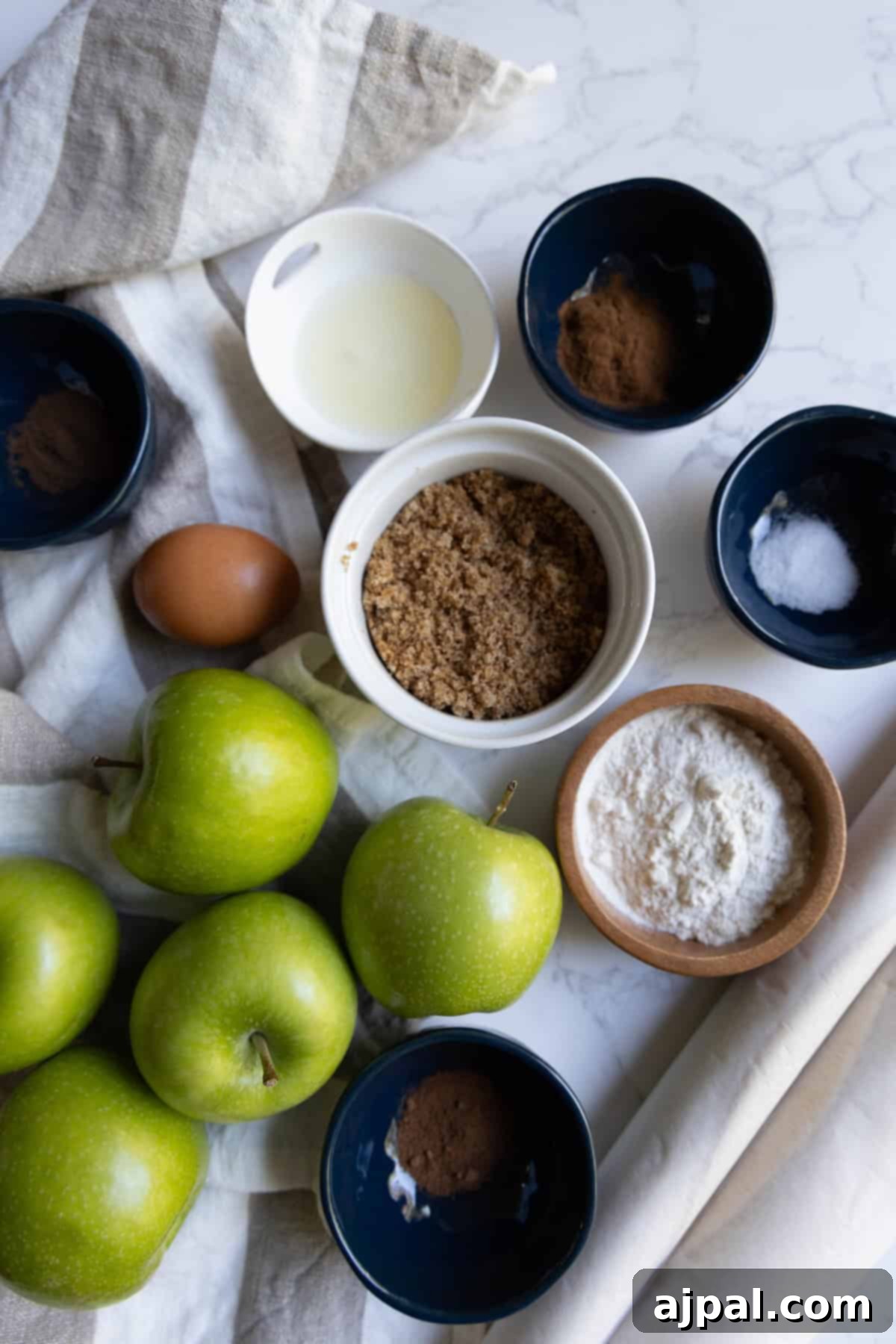 Heirloom Apple Pie 7 Overhead view of baking ingredients for apple pie, including green apples, brown sugar, flour, an egg, spices, baking powder, and a small bowl of liquid on a white surface with a striped towel.