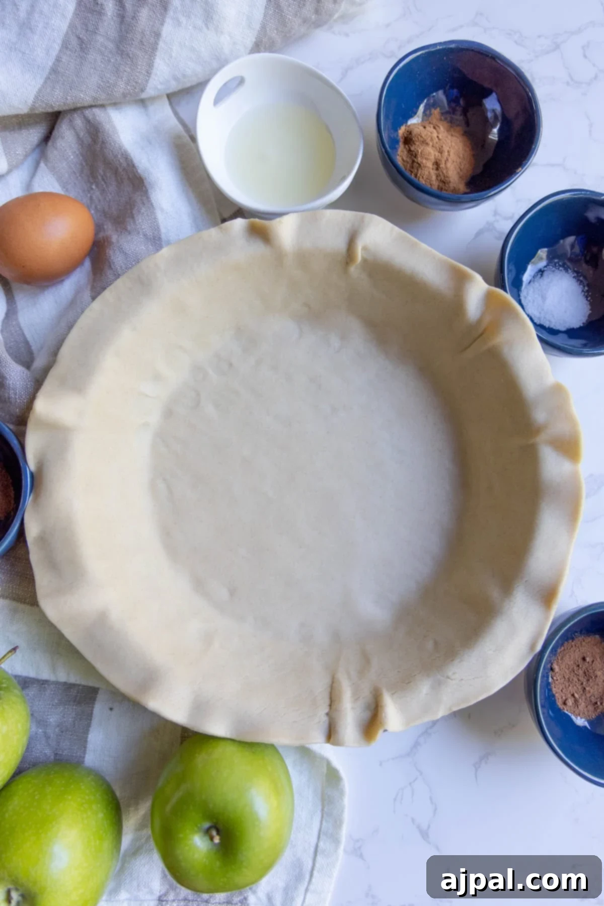 Heirloom Apple Pie 8 Unbaked pie crust in a pie dish surrounded by bowls of ingredients, a whole egg, and green apples on a white surface—perfect for creating a classic apple pie.