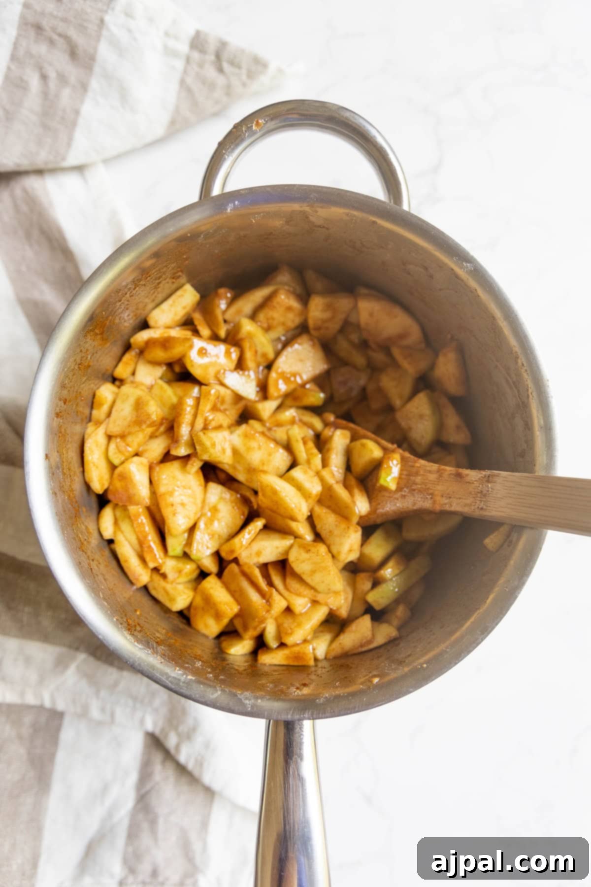 Heirloom Apple Pie 9 Sliced apples mixed with apple pie spices in a saucepan, stirred with a wooden spoon, on a light surface with a striped cloth nearby.