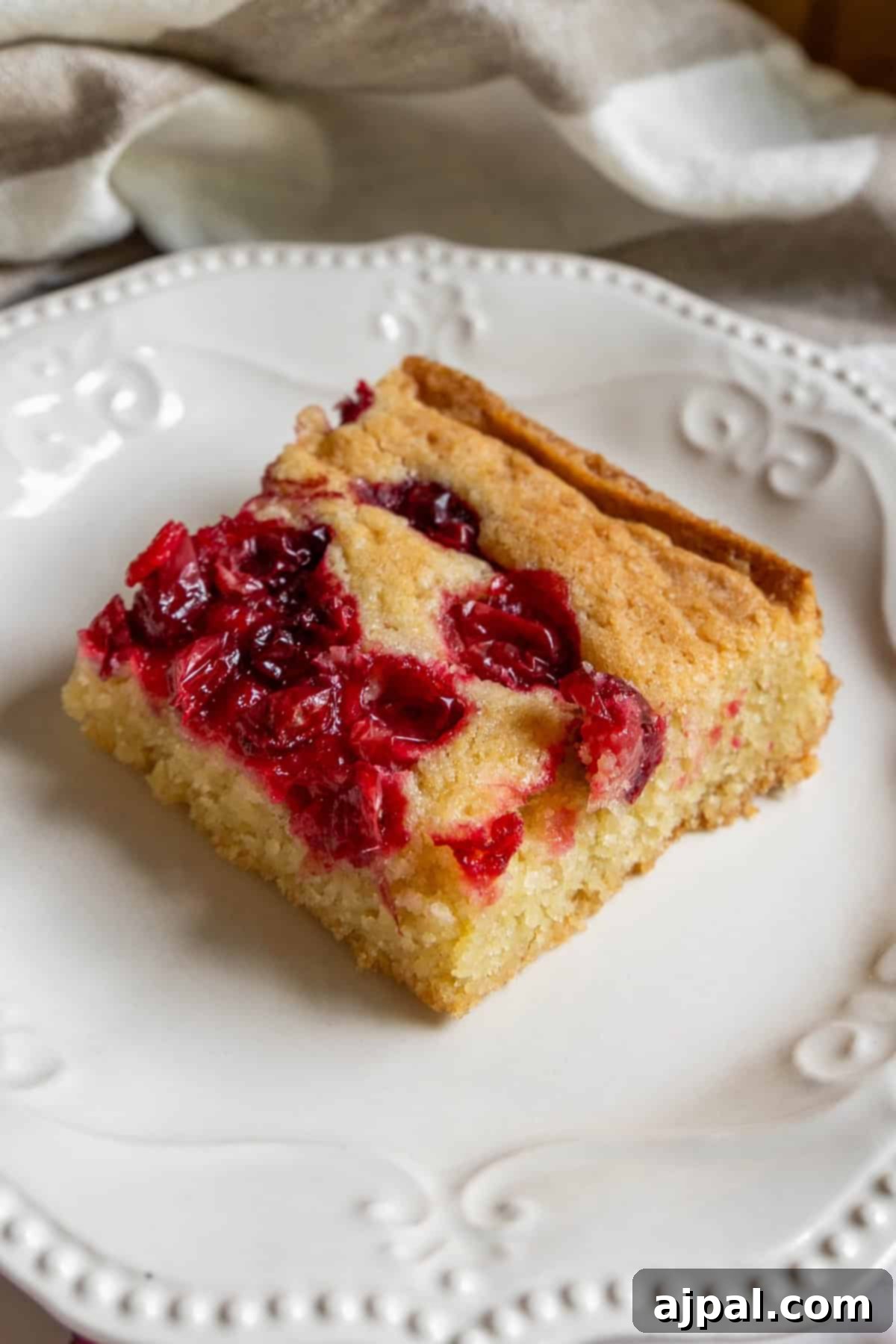 A square slice of golden cranberry bar cake, adorned with vibrant red cranberries, elegantly presented on a white decorative plate.