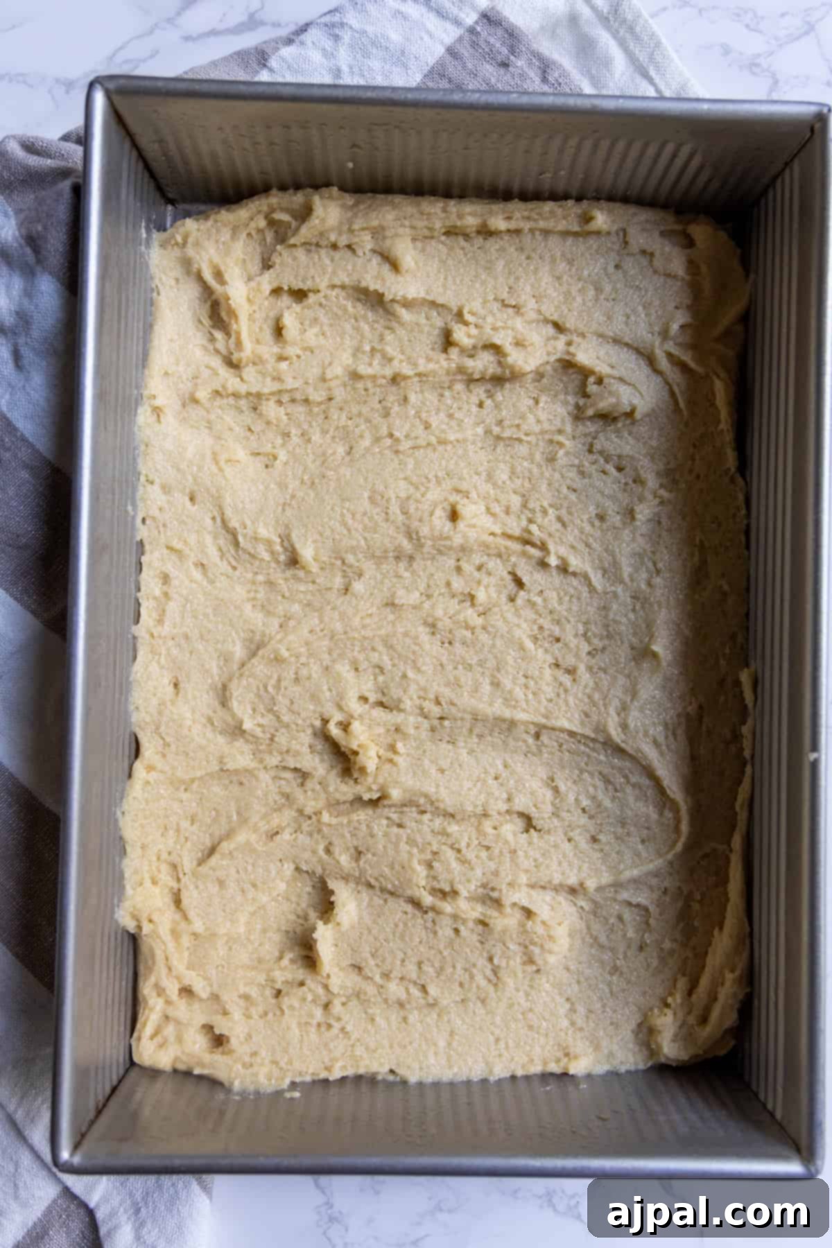 Rectangular metal baking pan filled with an even layer of raw cranberry bars dough, resting on a folded kitchen towel, ready for the next step.