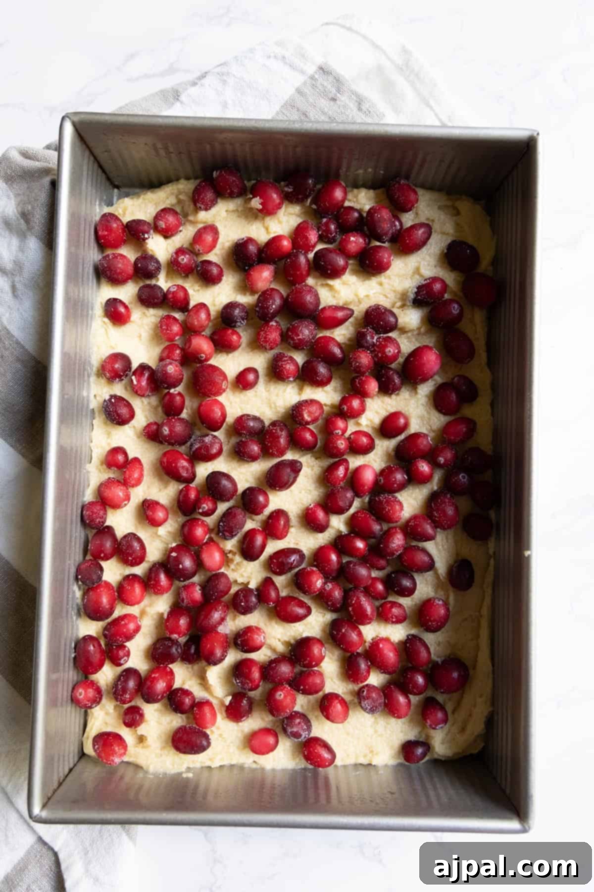A rectangular metal baking pan filled with unbaked cranberry bars dough topped with a generous layer of fresh red cranberries, ready for baking.