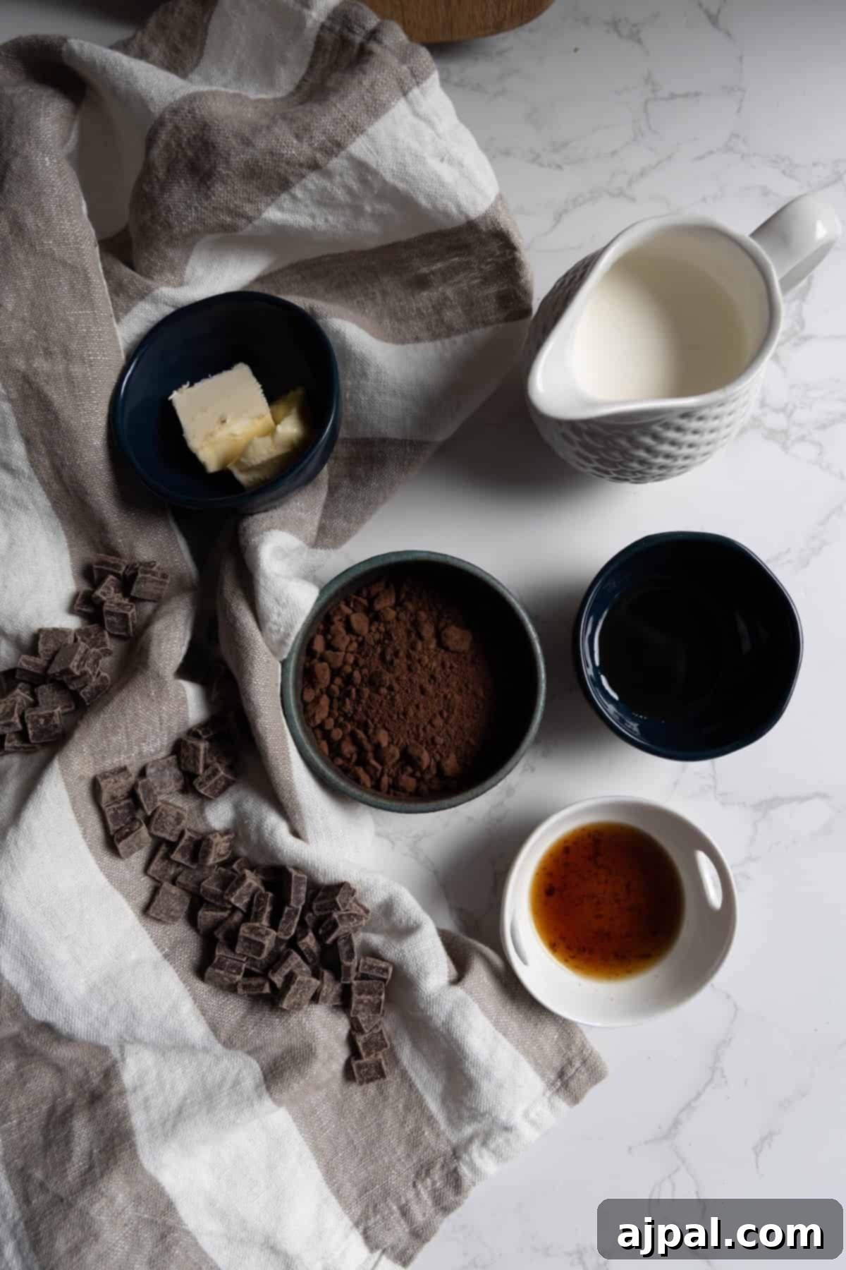 Several small bowls with butter, cocoa powder, cream, chocolate chunks, water, and vanilla extract—perfect ingredients for chocolate truffles—are arranged on a striped cloth on a white marble surface.