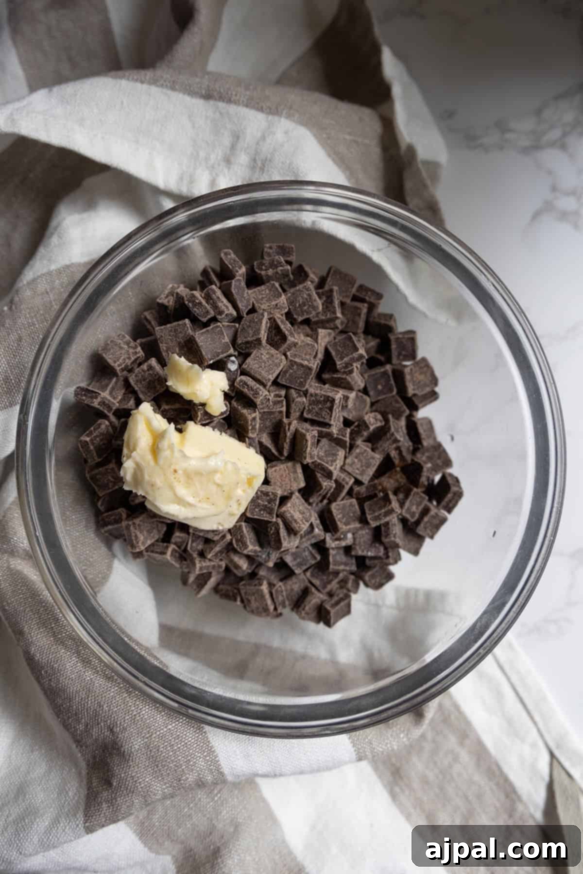 A glass bowl filled with chocolate chunks and a dollop of butter, perfect for making rich chocolate truffles, sits atop a striped cloth.