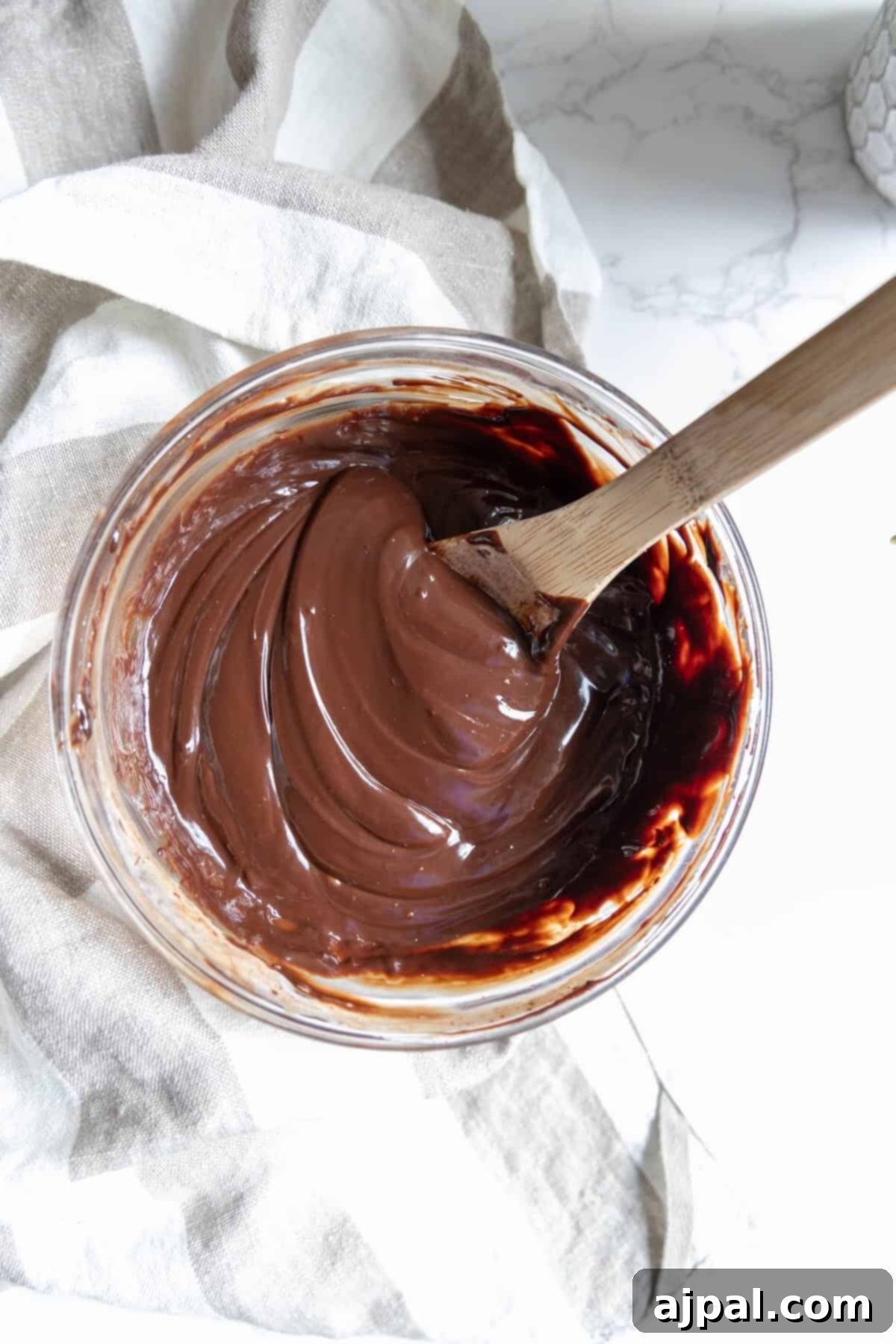 A glass bowl of smooth, glossy chocolate mixture for chocolate truffles sits with a wooden spoon resting inside, placed on a striped cloth.