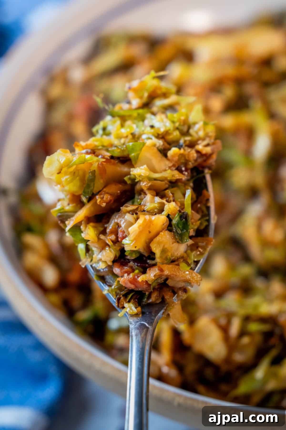 A close-up of a spoonful of cooked, shredded Brussels sprouts mixed with small pieces of crispy pancetta and seasonings, held above a bowl of the same savory dish.