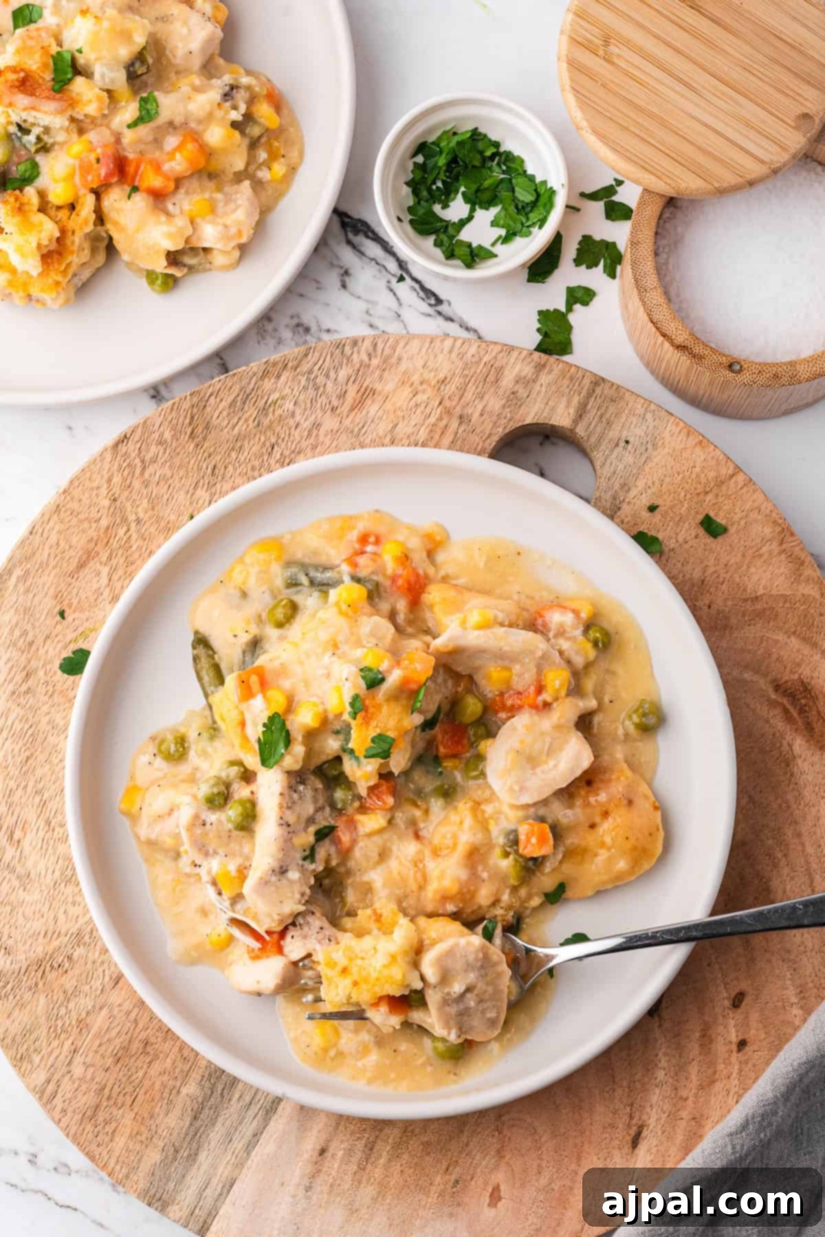 Simple Savory Chicken Bakes 2 A plate of creamy chicken pot pie casserole with vegetables rests on a wooden board, garnished with parsley, beside a small bowl of herbs and an open salt container. This savory dish is topped with a golden crust, revealing tender chicken and mixed vegetables in a rich sauce beneath.