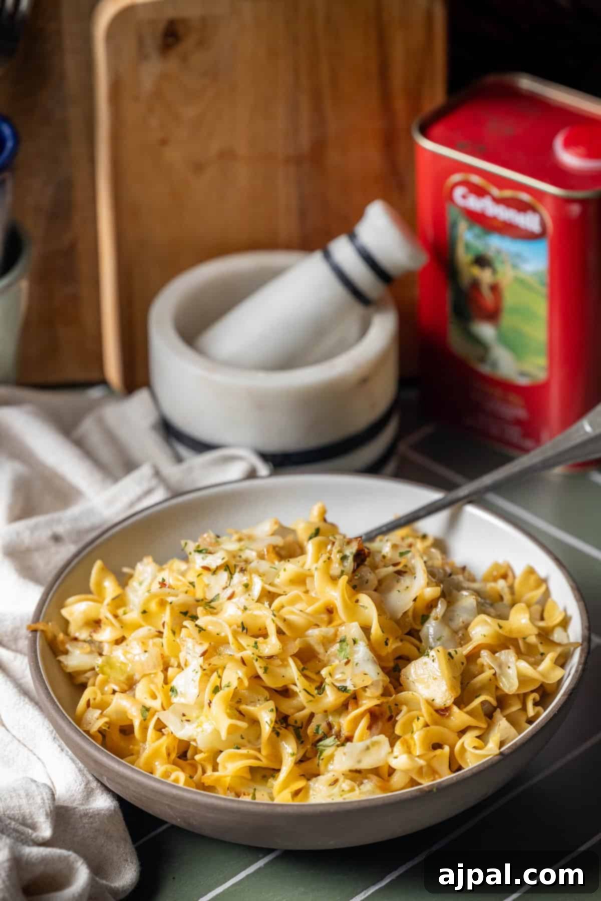Homestyle Cabbage Noodles 5 A bowl of egg noodles with sautéed onions and cabbage, topped with herbs, sits on a table with a mortar and pestle and a red food tin in the background.