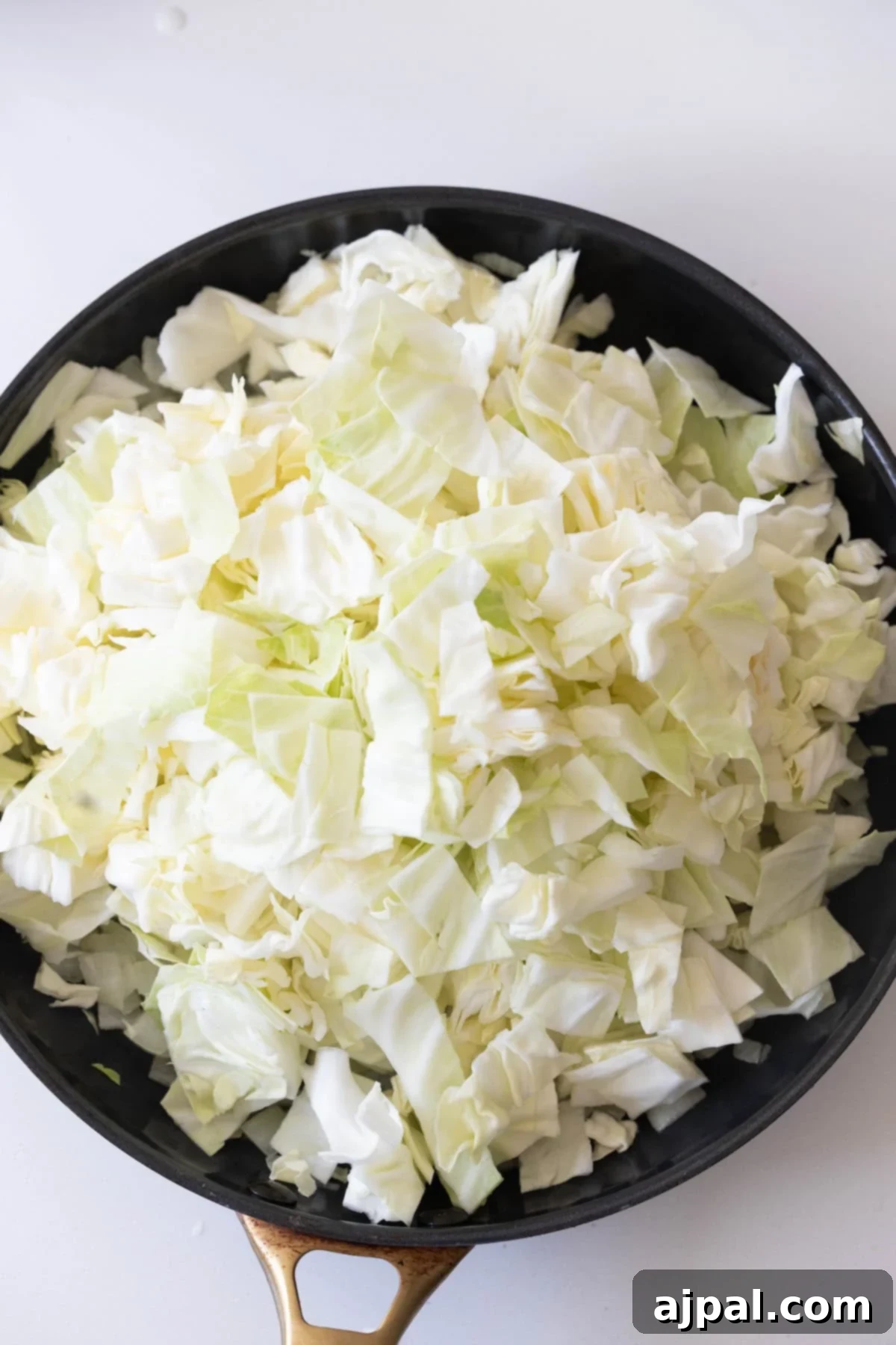 Homestyle Cabbage Noodles 7 Chopped raw cabbage piled in a black skillet on a white surface, ready to be cooked for a classic cabbage and noodles dish.