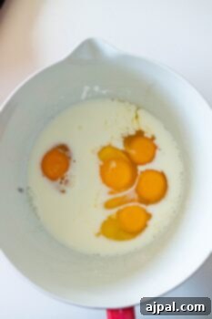 A white mixing bowl contains six raw eggs and milk, ready to be whisked together as the base for a classic corn pudding recipe, all set on a clean white surface.