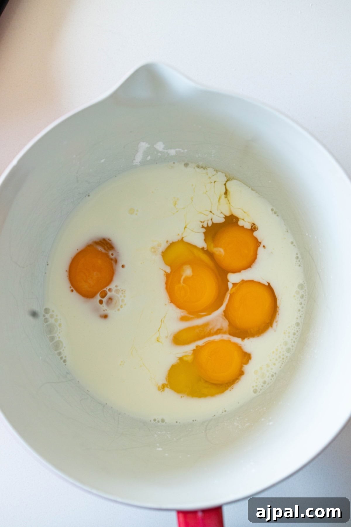 In a white mixing bowl, raw eggs and milk are whisked together, forming the creamy base for the classic corn pudding recipe on a pristine white surface.