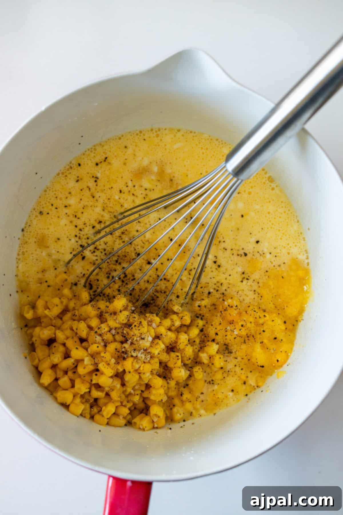 A whisk blends beaten eggs, sweet corn kernels, and ground black pepper in a white bowl, preparing the mixture for an easy and flavorful corn pudding.