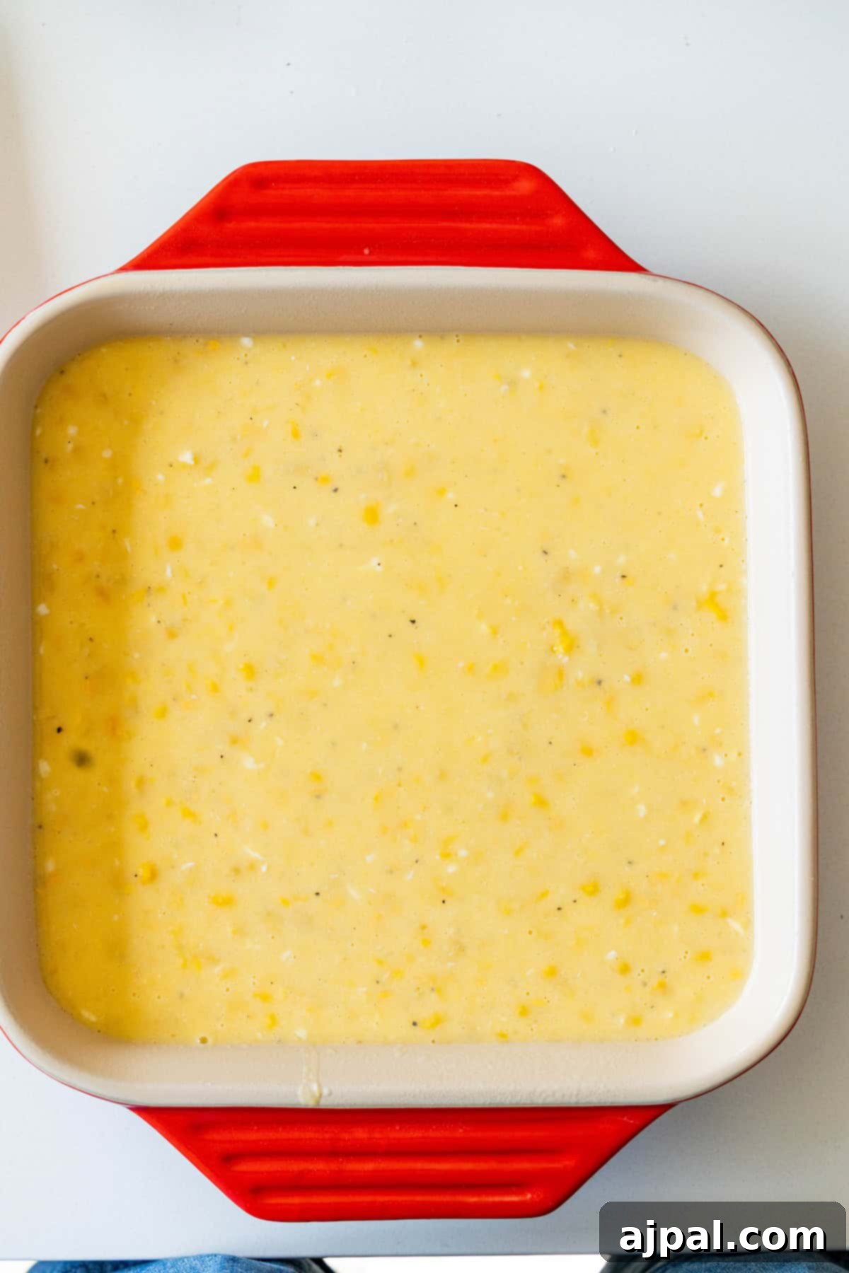 Uncooked corn pudding batter fills a vibrant red and white square baking dish, ready for the oven to transform it into a savory holiday side.