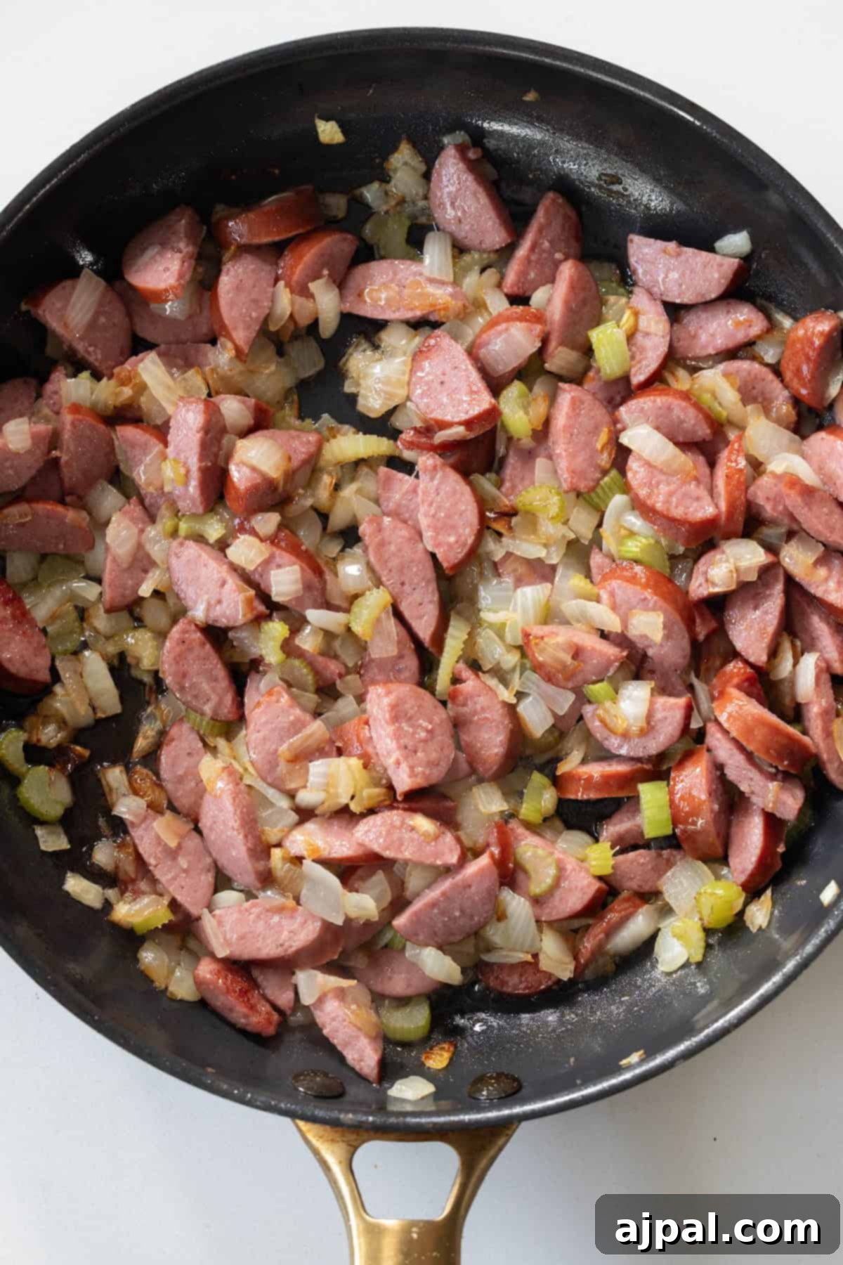 Sliced Polish Kielbasa, chopped onions, and celery are sautéing in a black skillet with a gold handle—the perfect starting point for a hearty and flavorful kielbasa stuffing.