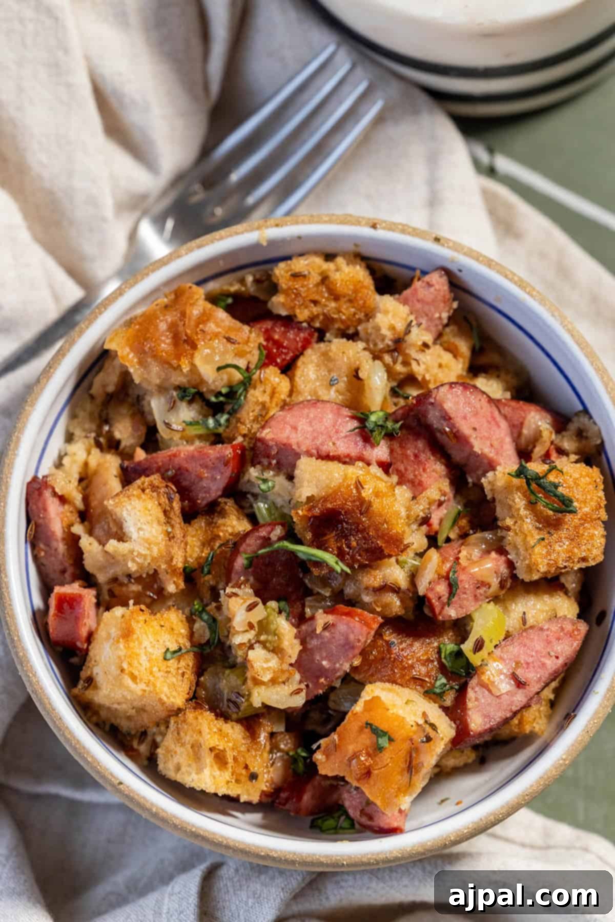 A tempting bowl of bread stuffing, perfectly mixed with succulent Polish Kielbasa slices, aromatic herbs, and colorful diced vegetables, elegantly presented on a light cloth with a fork on the side.