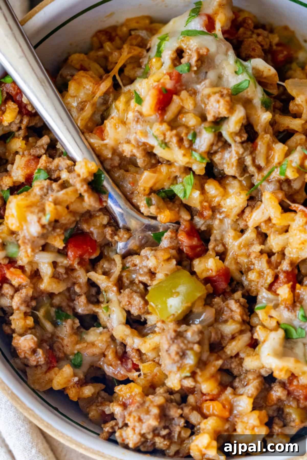 Fiery One-Pot Taco Skillet 2 Close-up of a bowl of cheesy ground beef and rice casserole with tomatoes, bell peppers, and herbs—a savory One Pot Spicy Taco Rice Skillet—served with a spoon.