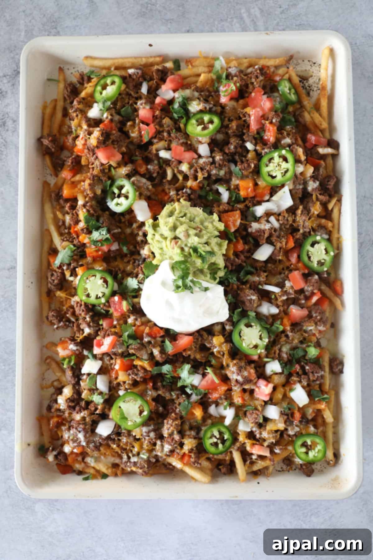 A baking sheet filled with loaded nacho fries topped with ground beef, melted cheese, diced tomatoes, onions, jalapeños, cilantro, guacamole, and sour cream for the ultimate snack.