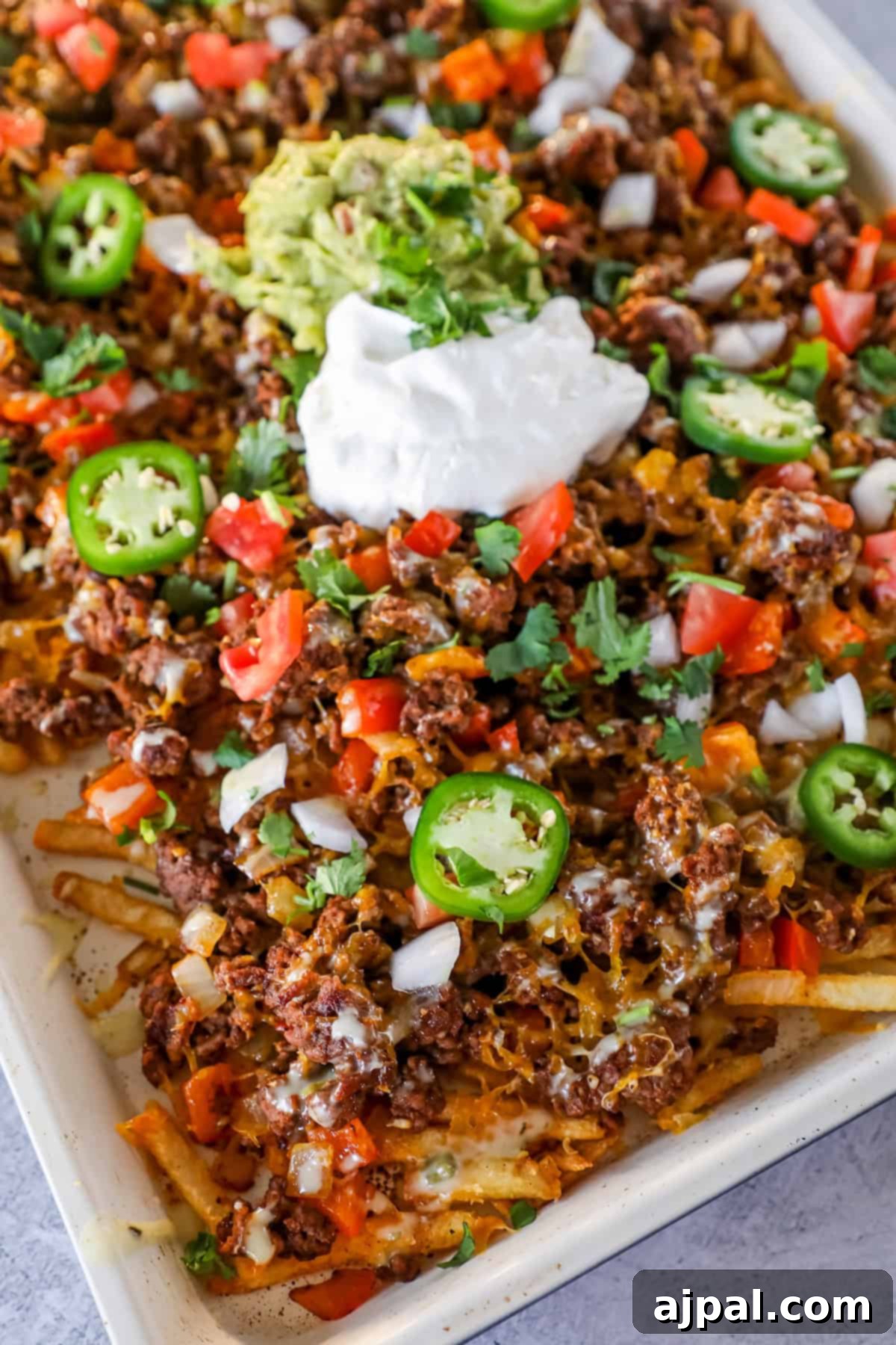 A sheet pan topped with loaded nacho fries—crispy French Fry Nachos piled high with ground beef, melted cheese, diced tomatoes, onions, jalapeño slices, cilantro, guacamole, and a dollop of sour cream.