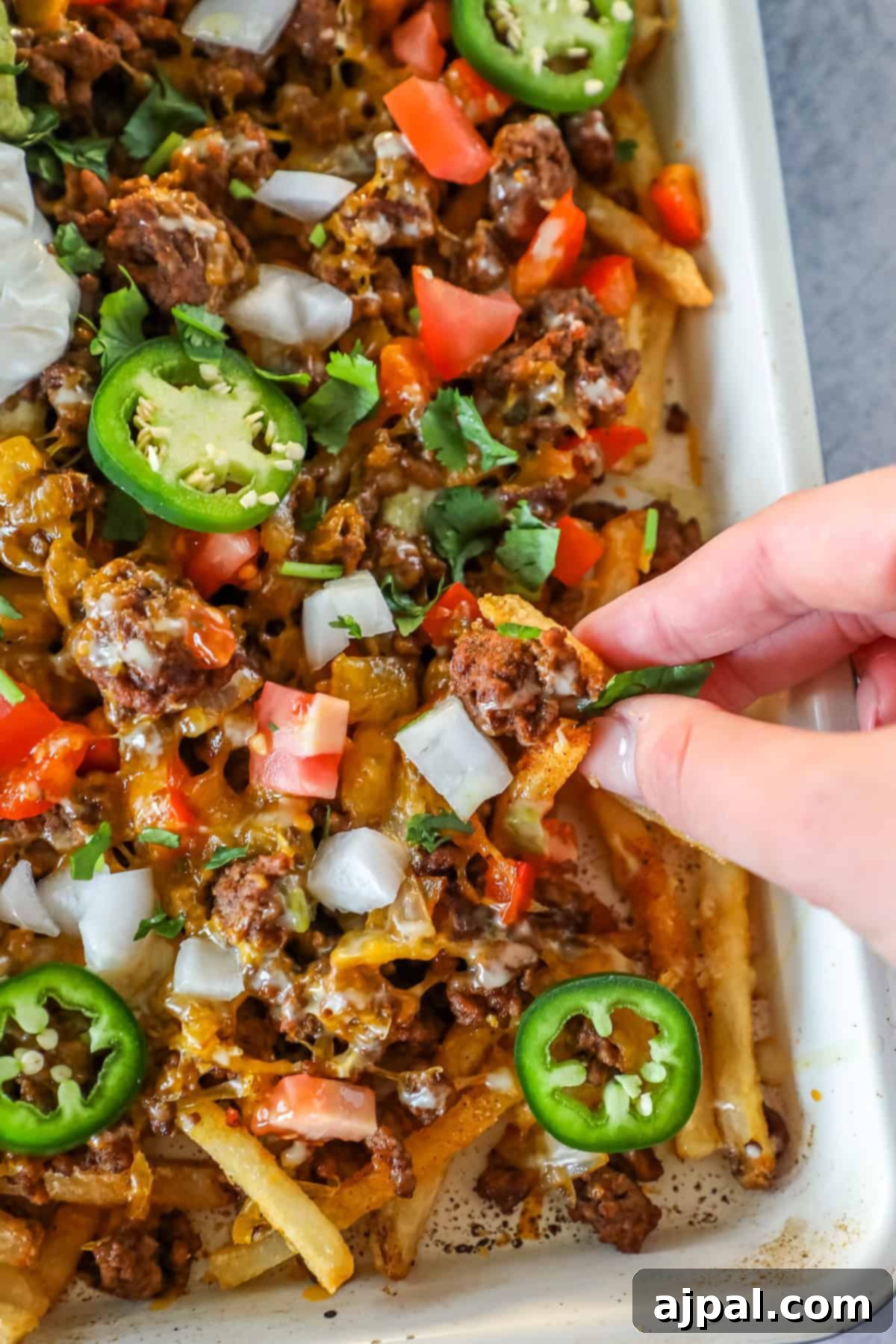 A hand picks up loaded nacho fries, topped with ground beef, melted cheese, diced tomatoes, onions, jalapeños, and cilantro from a tray—perfect for those craving easy nacho fries packed with bold flavor.
