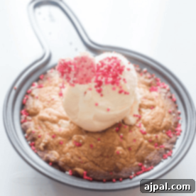 A baked dessert of brown butter blondies in a round pan topped with a scoop of vanilla ice cream, pink and red sprinkles, and heart-shaped candies.