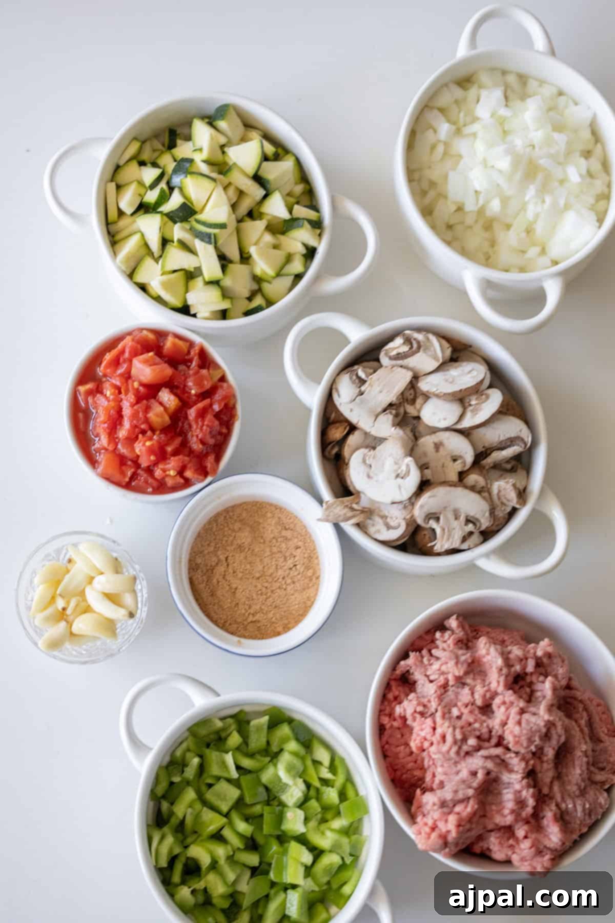 Loaded Cheesy Taco Skillet 3 Several bowls filled with chopped zucchini, onions, tomatoes, mushrooms, green bell pepper, minced garlic, breadcrumbs, and ground meat are arranged on a white surface—perfect for preparing the Best Taco Skillet.