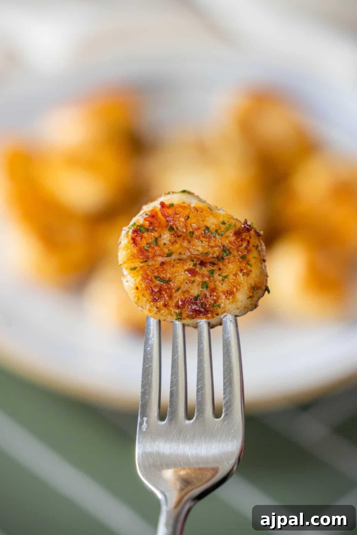 Caramelized Pan Seared Scallops 2 A cooked, seasoned Perfect Pan Seared Scallop is held on a fork, with more scallops blurred on a plate in the background.