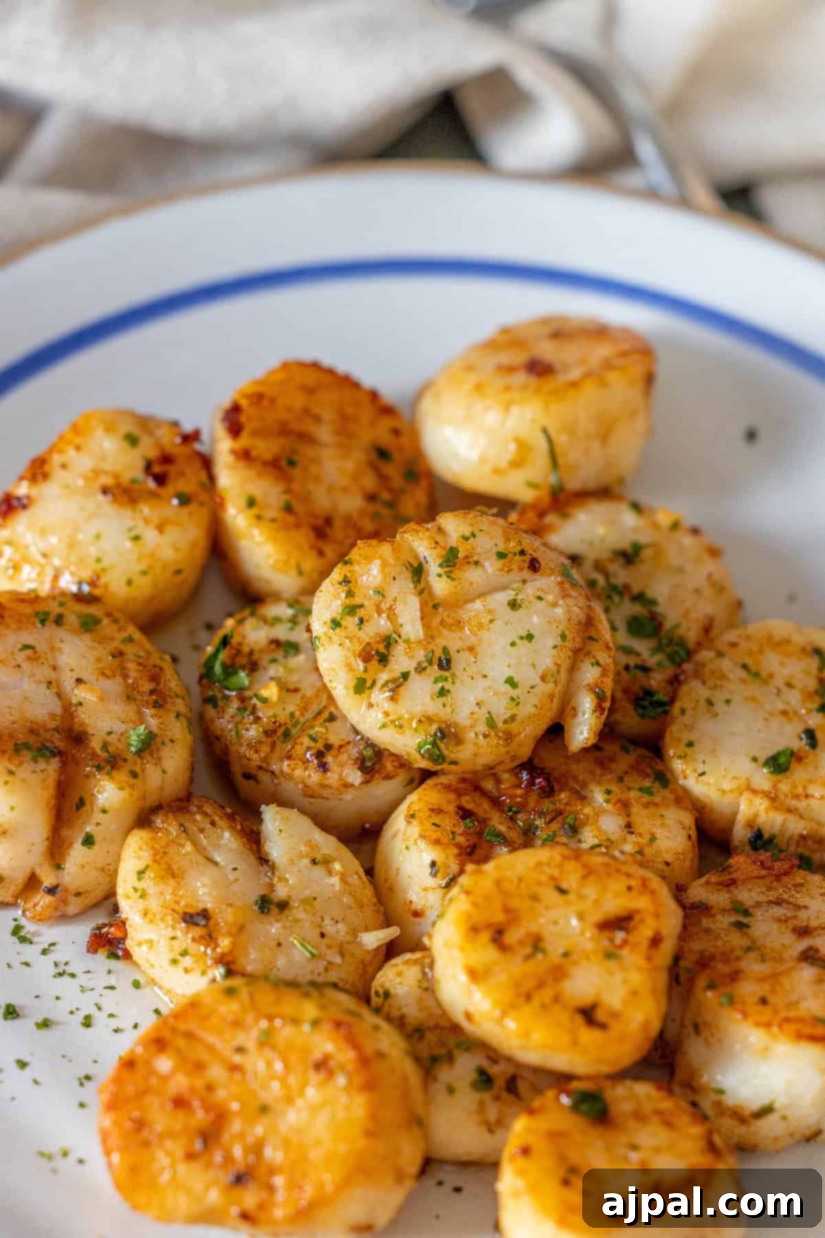 Caramelized Pan Seared Scallops 3 A white plate with Perfect Pan Seared Scallops, garnished with chopped herbs, set on a table with a fork and napkin in the background.