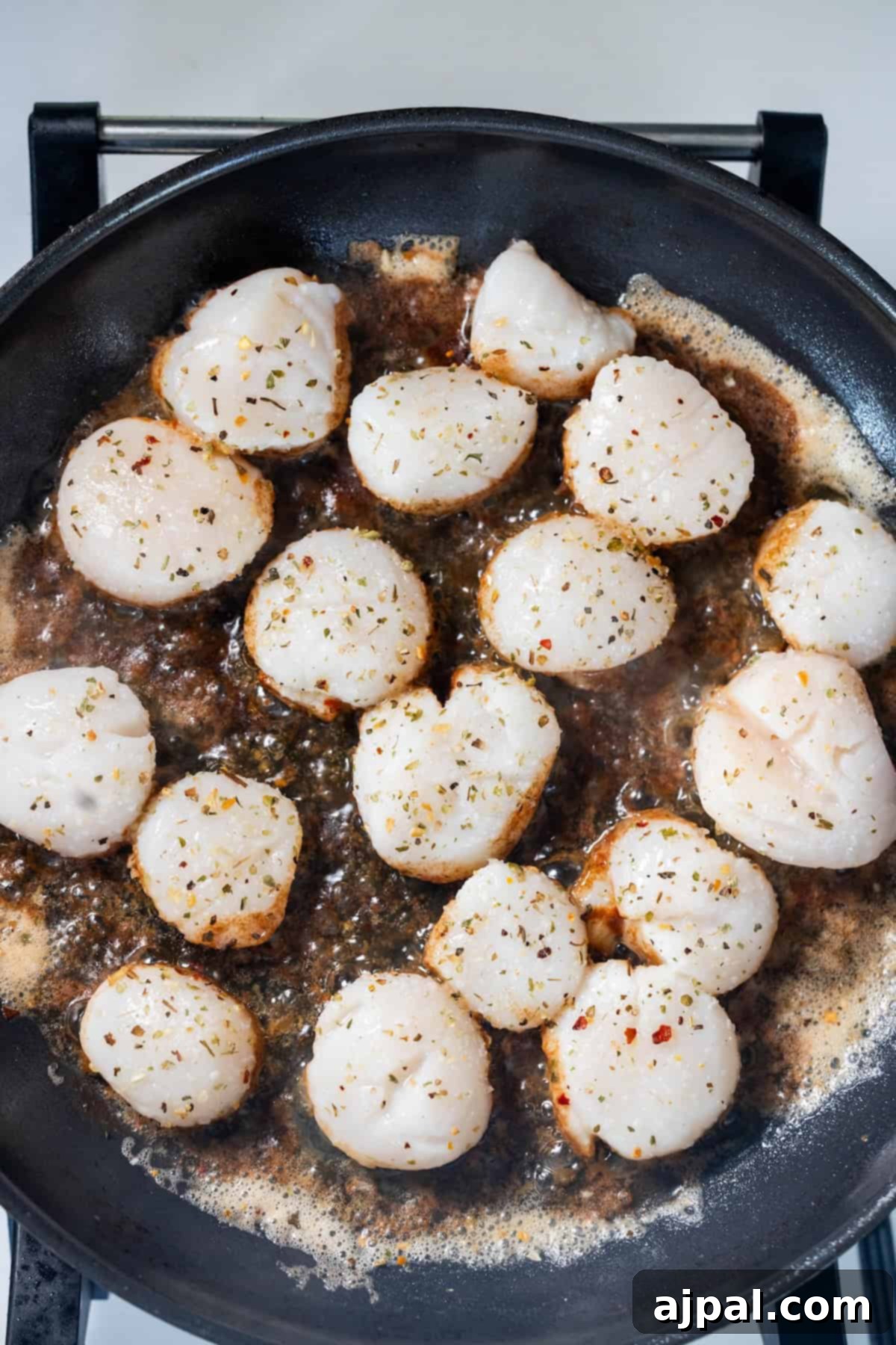 Caramelized Pan Seared Scallops 7 Perfect Pan Seared Scallops cooking in a frying pan with bubbling sauce, sprinkled with fresh herbs and spices.