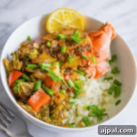 Paleo Salmon Taco Power Bowls 7 A Paleo bowl featuring grilled salmon atop cauliflower rice, complemented by sautéed vegetables, green onions, and a lemon slice, sits elegantly on a marble surface.