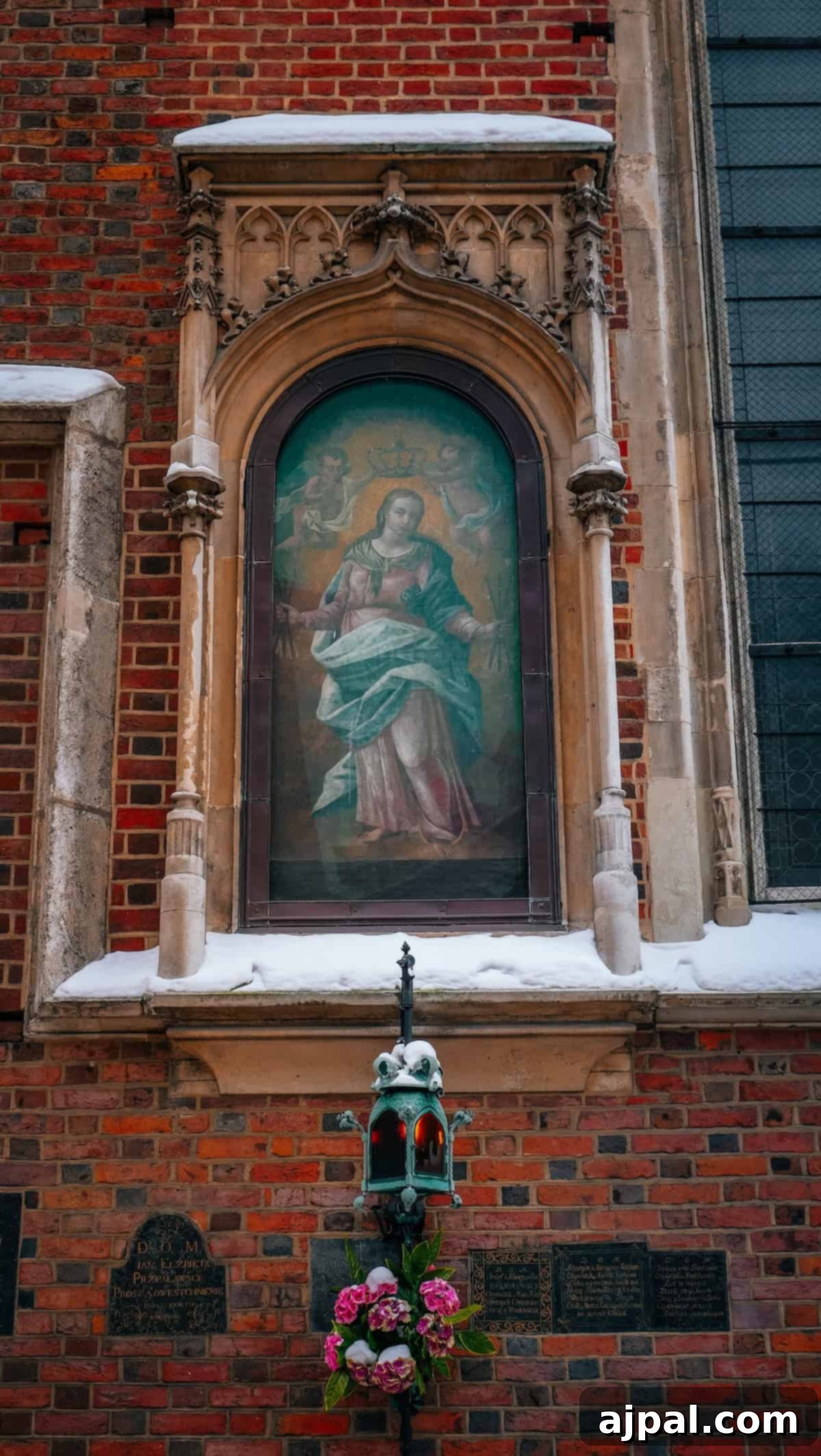 Krakow: Poland's Royal City 29 A religious painting in an ornate stone frame on a brick wall in Krakow, overlooking a small shrine with pink flowers and a lantern.