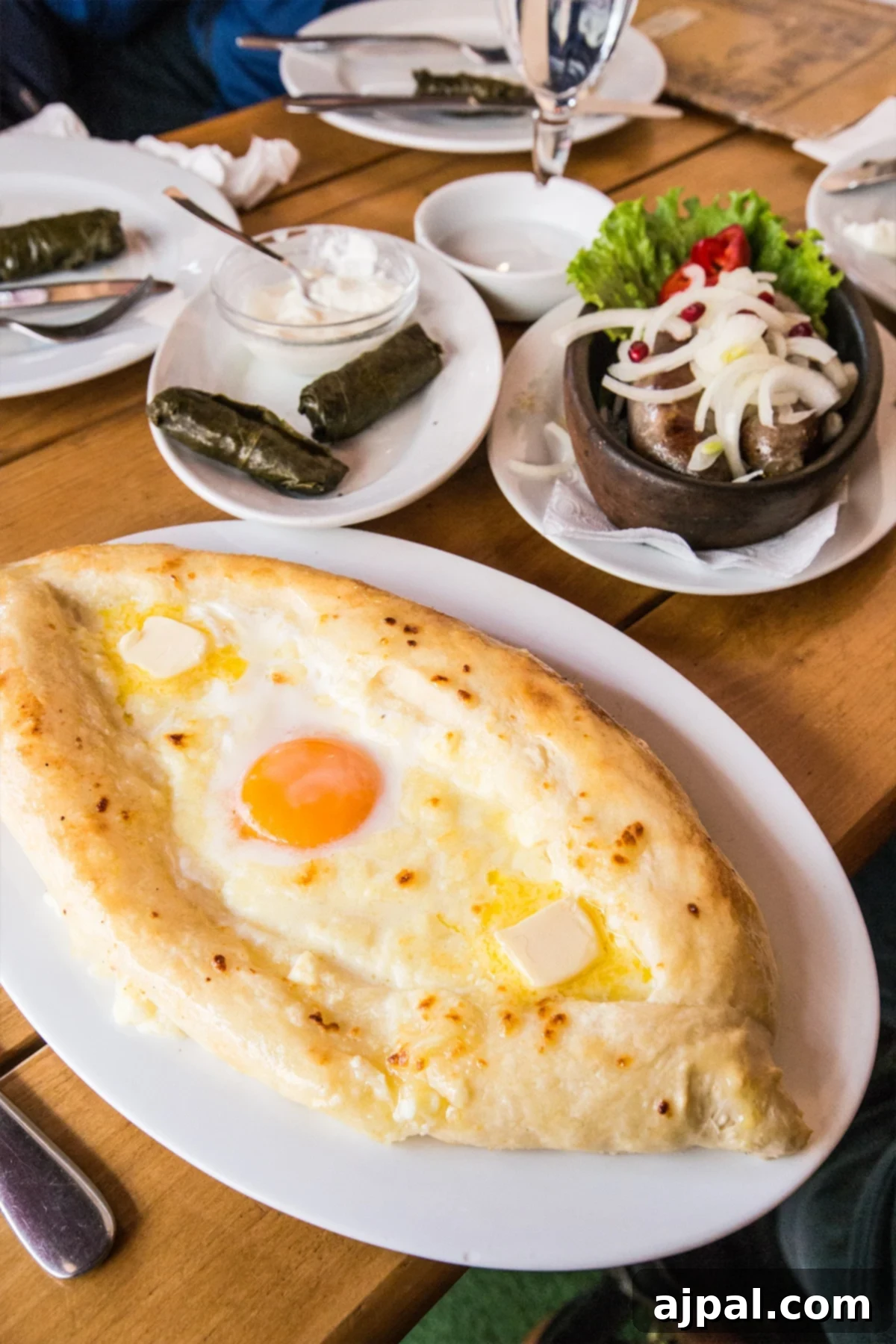A spread of traditional Georgian dishes including Khachapuri, sausages, and dolmades at a bustling cafe in Tbilisi.