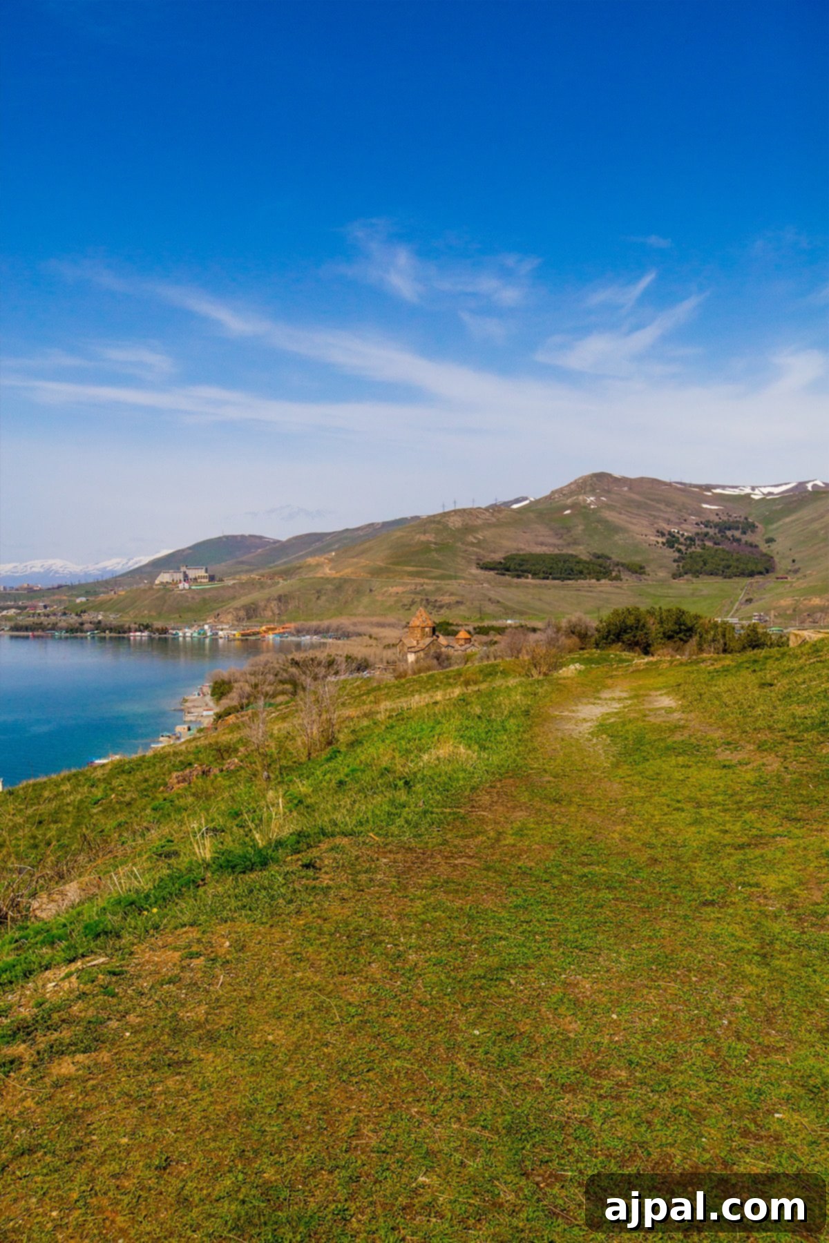 Lush green grassy fields stretching towards the serene Lake Sevan, with the majestic Sevanavank Monastery Ruins visible in the background, Armenia.