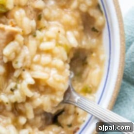 A close-up of a bowl of creamy risotto with pieces of chicken and herbs, being scooped with a fork. This easy chicken risotto recipe is perfect for one-pot cooking.