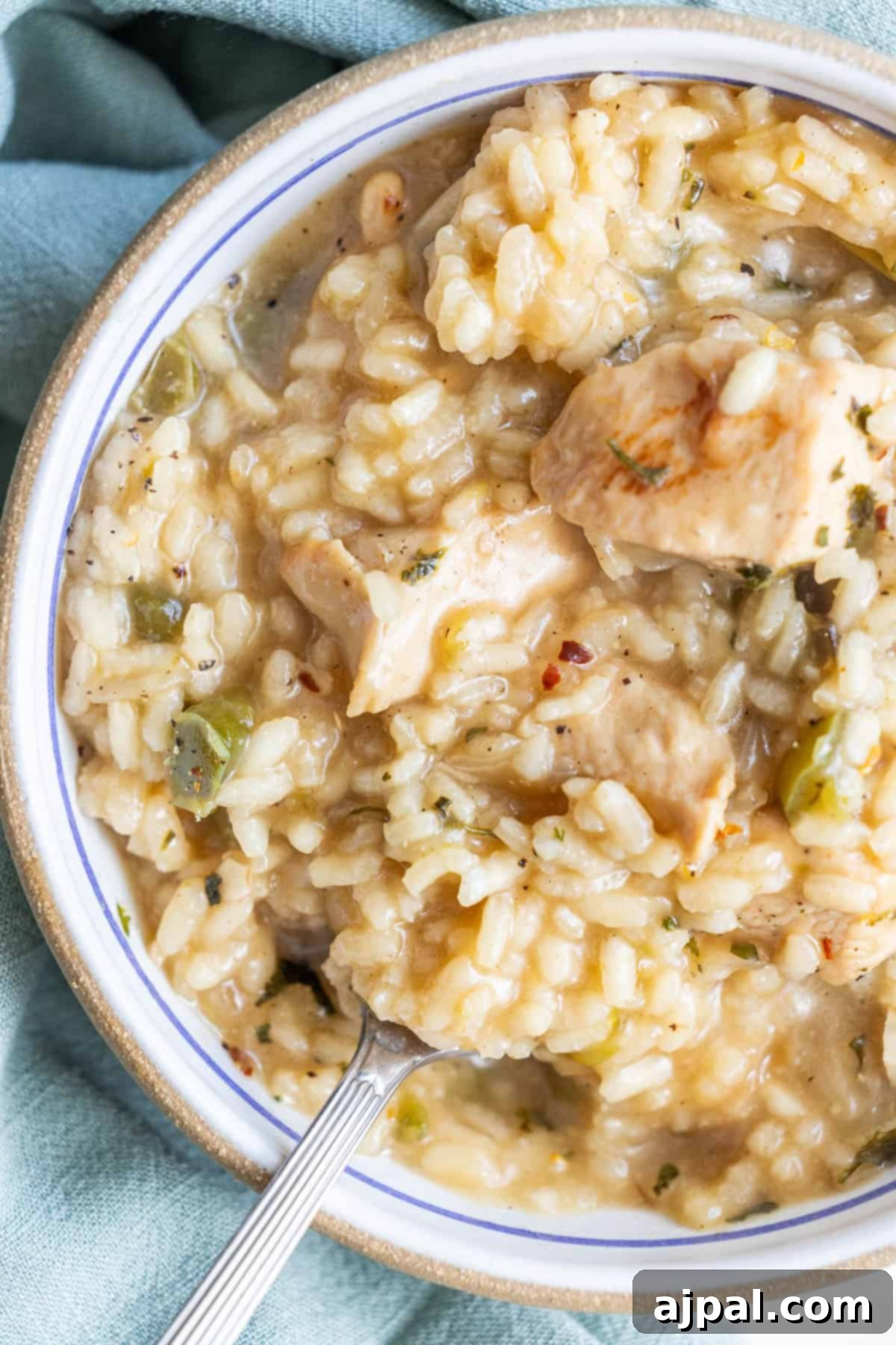 A close-up of a bowl of creamy one pot chicken risotto, featuring tender chunks of chicken, rice, and green vegetables, with a spoon partially dipped in the dish.