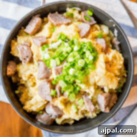 Hearty Cheesy Sausage Potato and Cabbage Bake 7 A bowl filled with mashed potatoes topped with sliced sausage and chopped green onions. Channeling a cheesy colcannon recipe, this hearty dish rests on a striped cloth, with a fork ready to dive in.