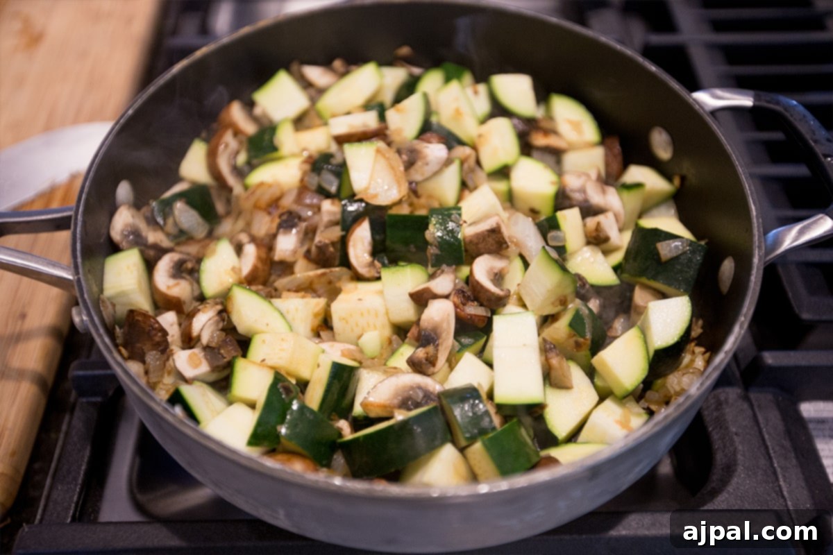 Chopped zucchini and mushrooms sizzling in a large pan, beginning to caramelize.