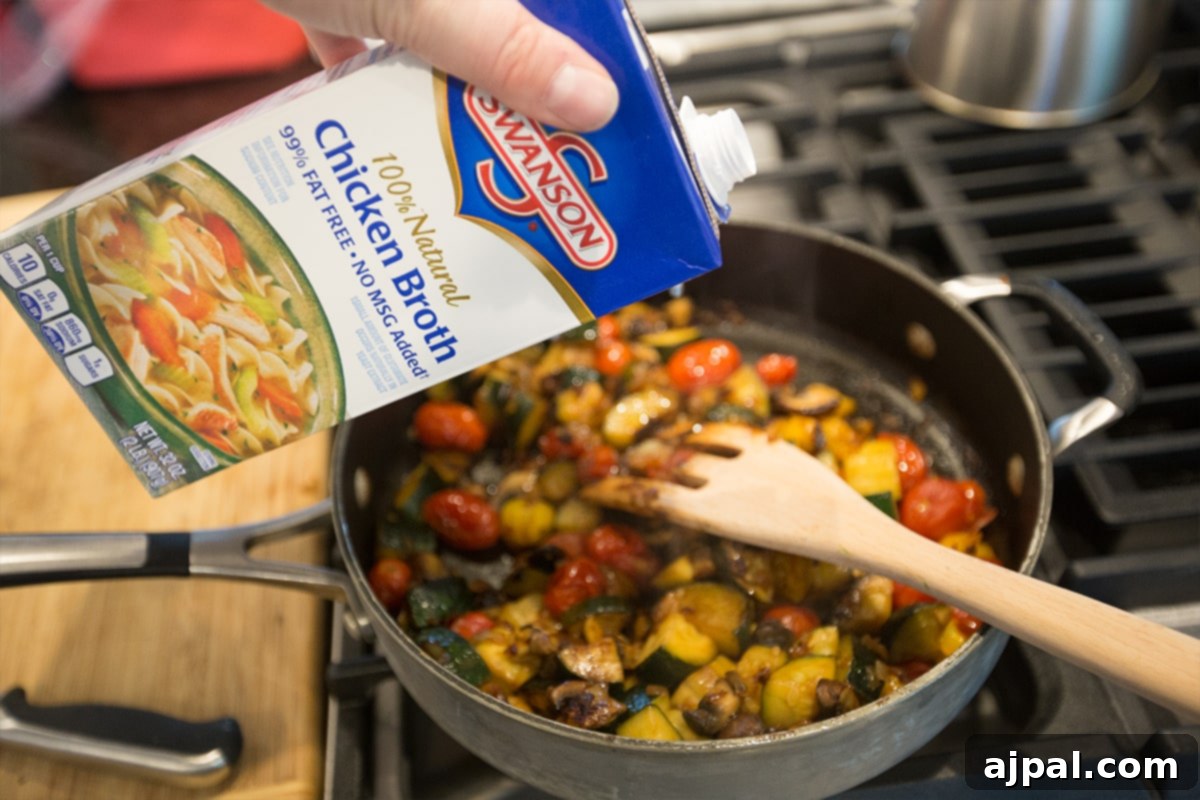 A pan with sautéed vegetables, with Swanson chicken broth being poured over them, ready for the couscous.