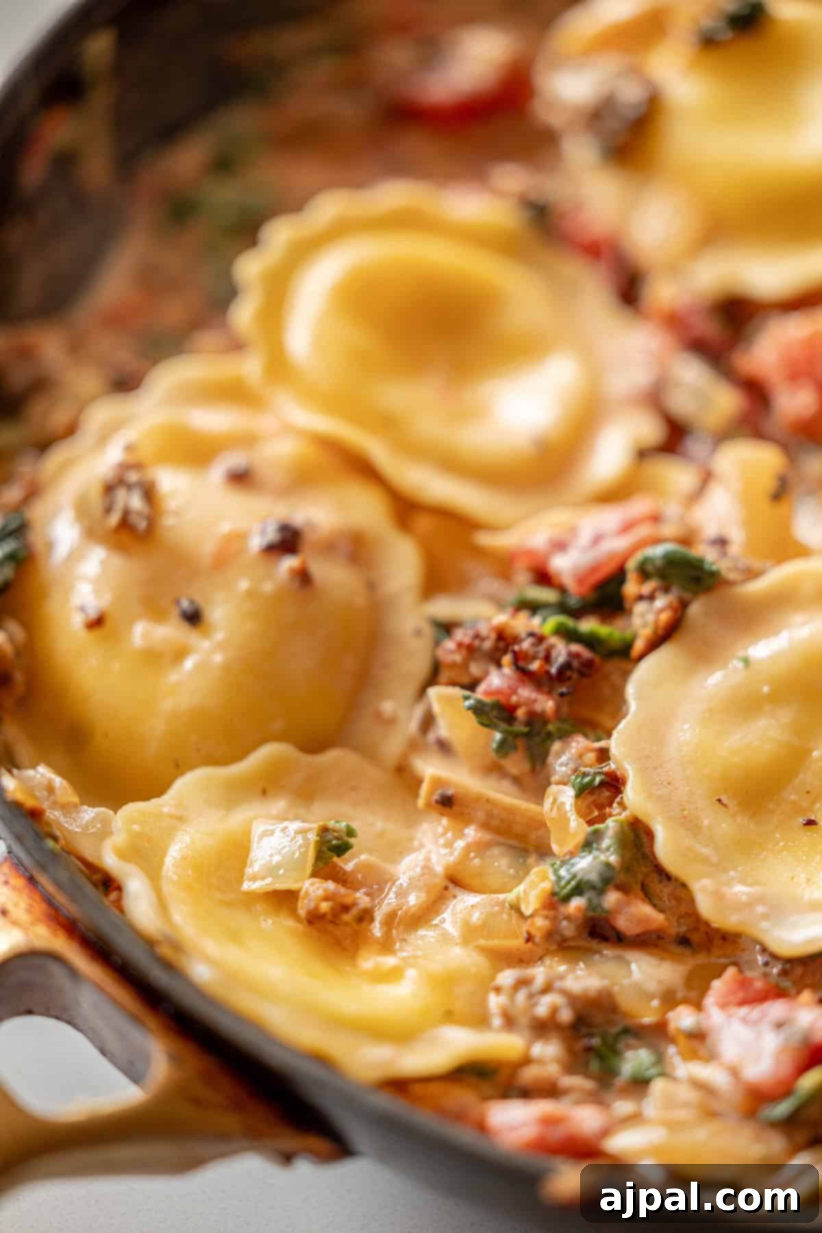 Creamy Tuscan Sausage Ravioli 11 A close-up of delicious tomato cream sauce ravioli in a skillet, generously mixed with spinach, sun-dried tomatoes, and onions for a rich and creamy blend.