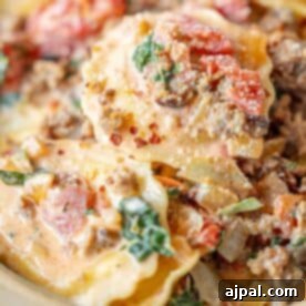 Close-up of ravioli with tomato spinach cream sauce, ground meat, fresh spinach, tomatoes, onions, and a creamy base, topped with grated cheese and red pepper flakes.