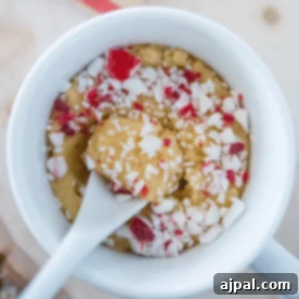 A white mug filled with Microwave Peppermint Cookie Dough Fudge for One, topped with crushed red and white peppermint candy, with a spoon inside the mug.