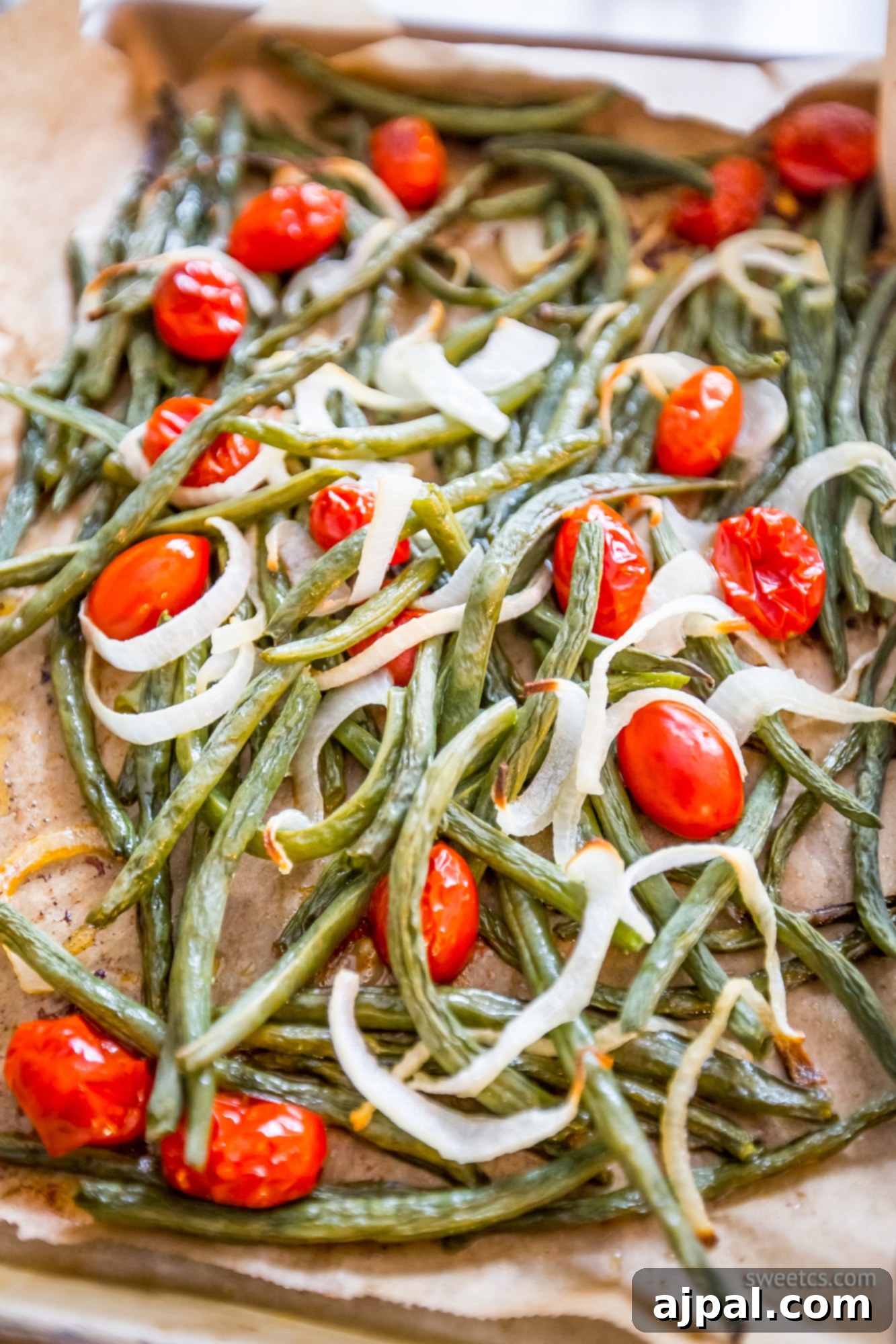 Close-up of roasted green beans and tomatoes, glistening with olive oil and seasoned, on a baking sheet, showing light browning.